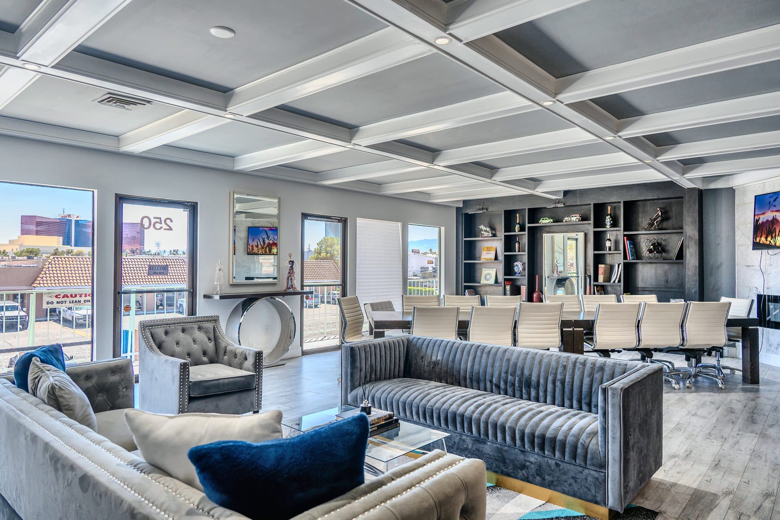 Executive lounge with a coffered ceiling, gray velvet sofas, a long boardroom table with cream chairs and a dark bookcase wall