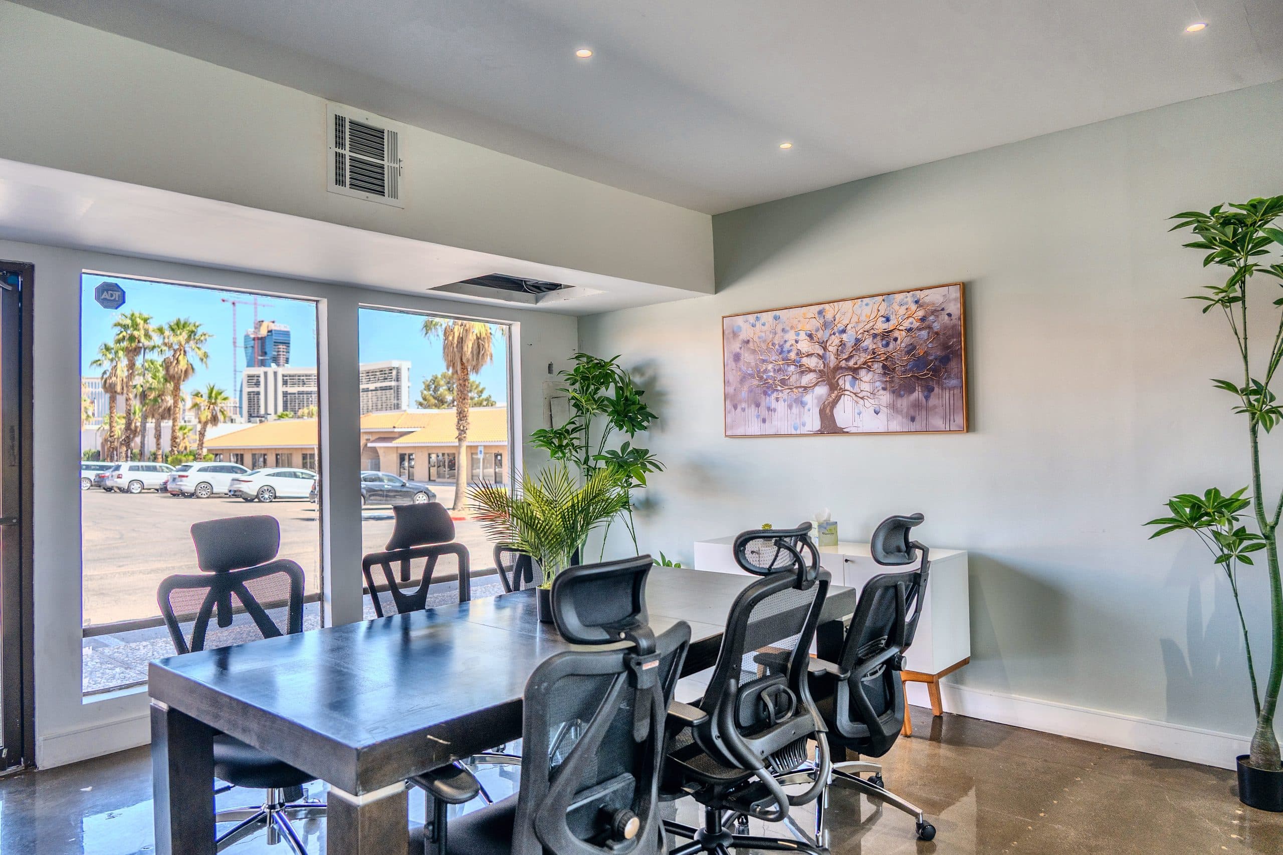 Meeting room with a dark wood table, black ergonomic chairs, a tree painting and windows onto palm trees and the Strip