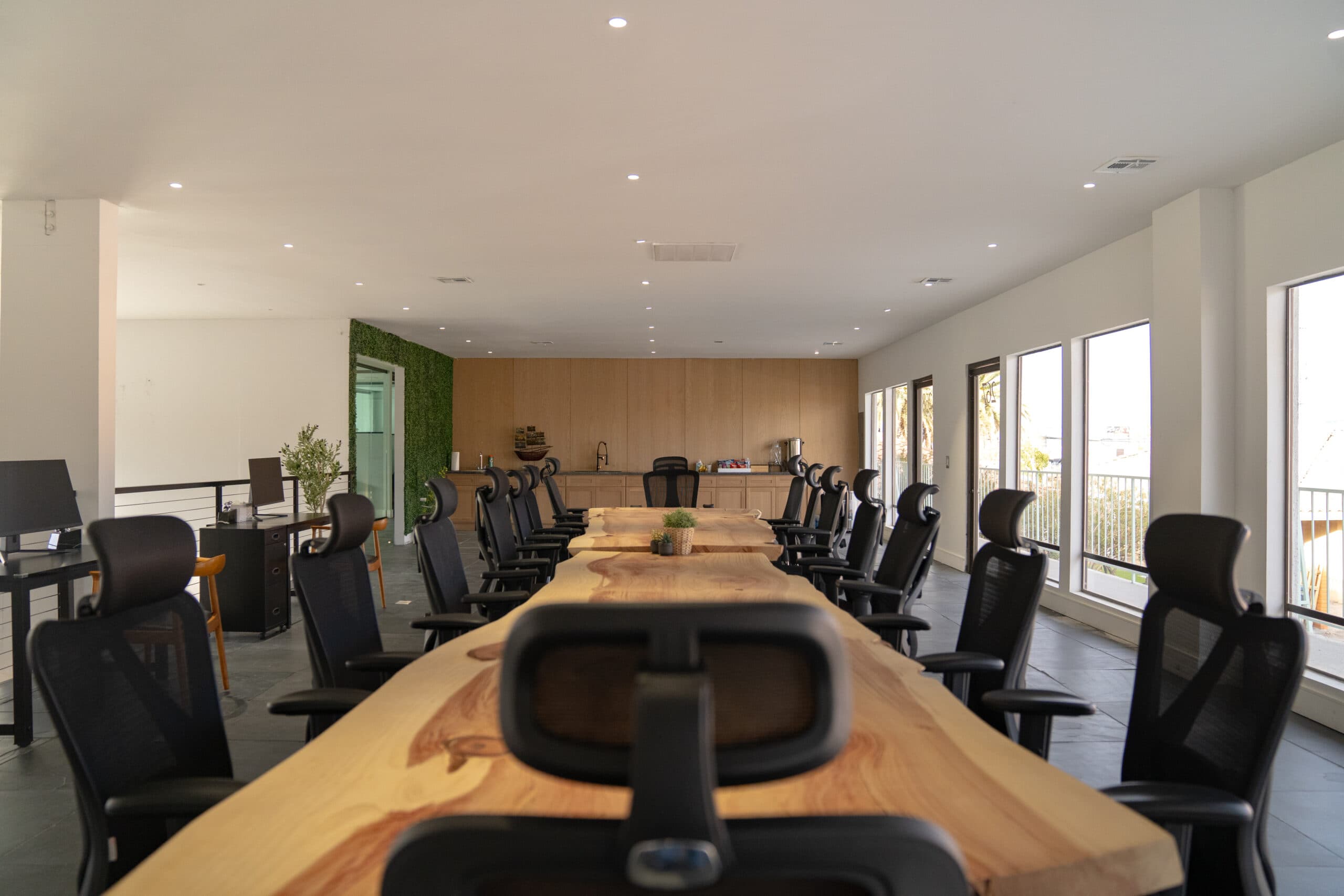 Mesa de reuniones larga de madera con bordes naturales, sillas de malla negra, una pequeña cocina con paneles de madera y una pared vegetal.