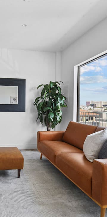 Cognac leather sofa and ottoman beside a potted plant, with a city view through the window