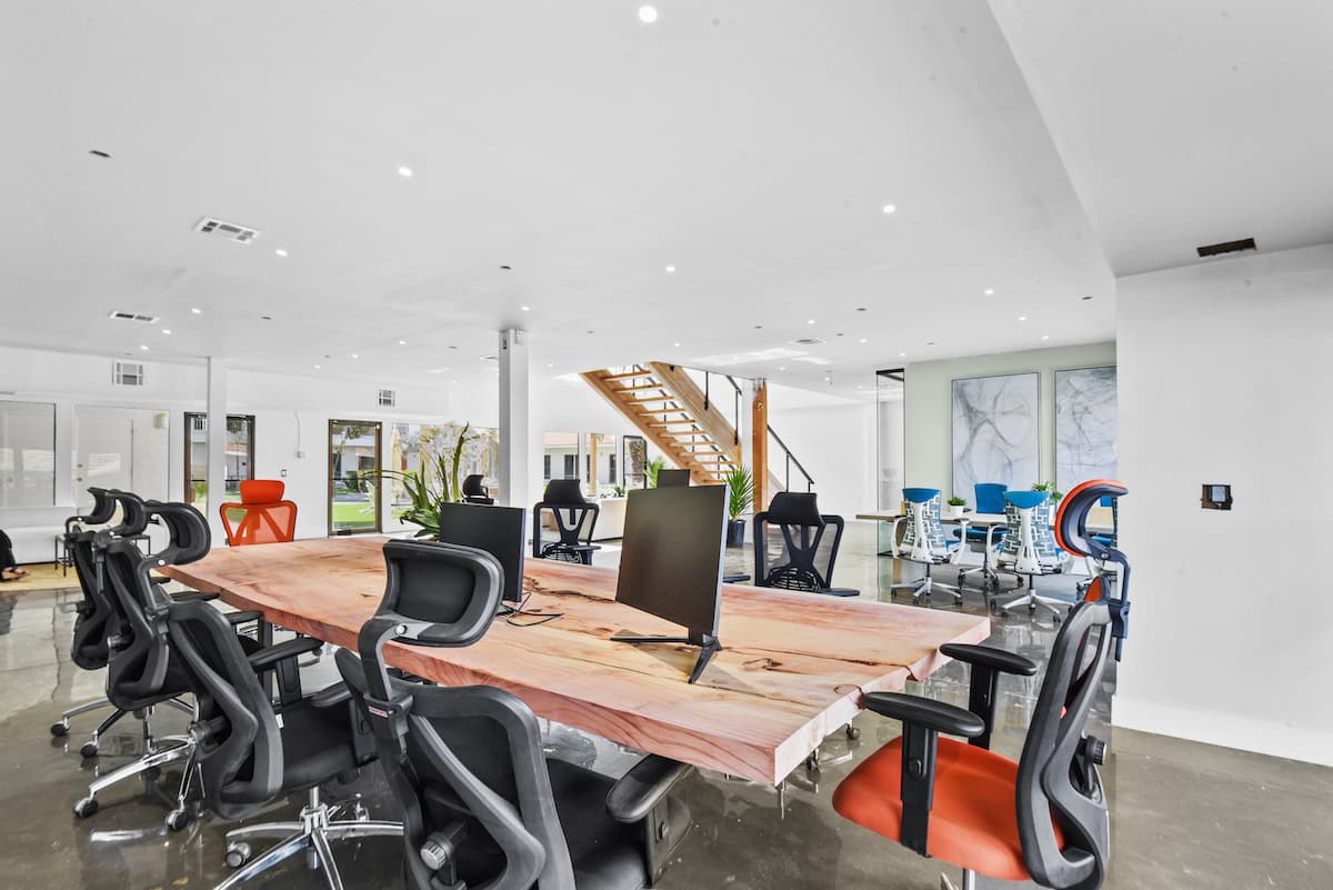 Open coworking floor with a live-edge slab desk, monitors and black and orange ergonomic chairs