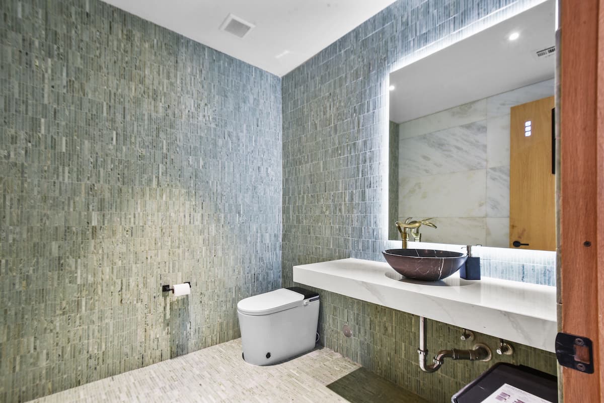 Aseo con paredes de azulejos de mosaico verde, un espejo retroiluminado, encimera de mármol y lavabo de piedra.