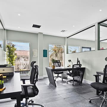 Private office with three black desks, ergonomic chairs and a printer, lit by windows with palm-tree views
