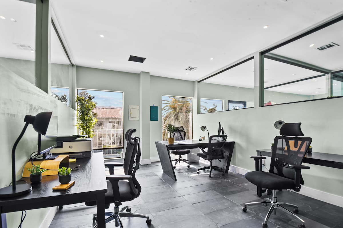 Private office with three black desks, ergonomic chairs and a printer, lit by windows with palm-tree views